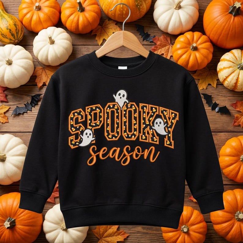 May include: A black long-sleeve sweatshirt featuring the text SPOOKY in orange and black candy corn patterned block letters and the word season in orange script. Three white ghost illustrations are positioned around the text.