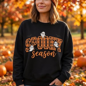 May include: A black crewneck sweatshirt featuring the text SPOOKY in an orange and black buffalo plaid pattern and the word season in orange script. The design includes three small white ghost illustrations and two black bat silhouettes.