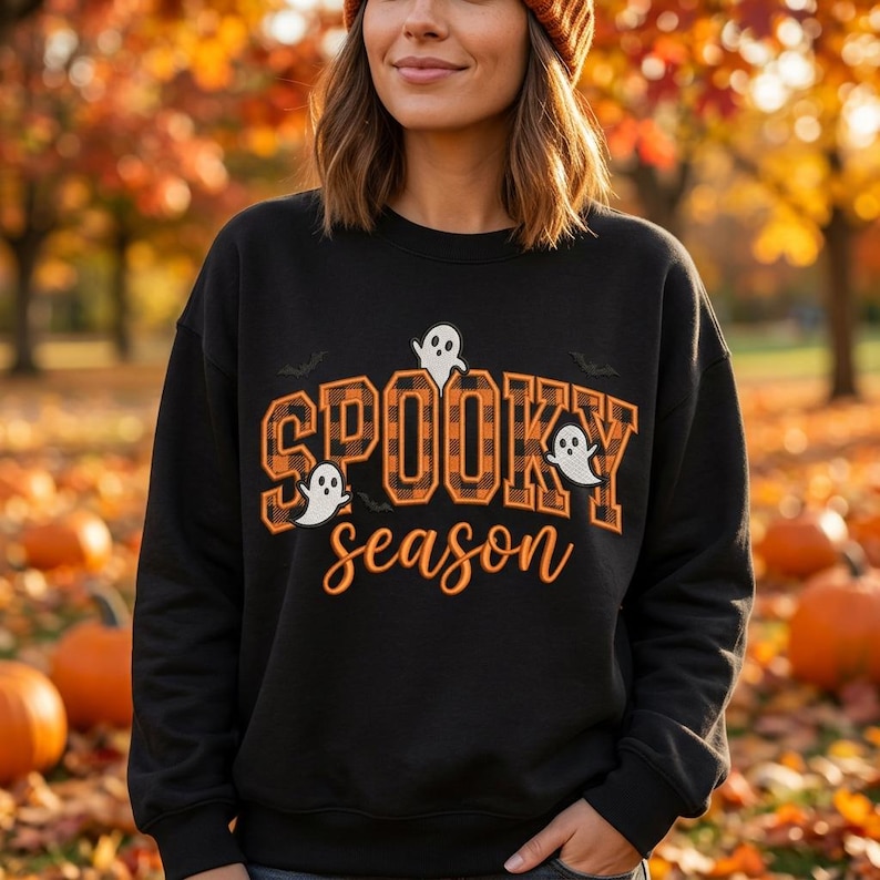 May include: A black crewneck sweatshirt featuring the text SPOOKY in an orange and black buffalo plaid pattern and the word season in orange script. The design includes three small white ghost illustrations and two black bat silhouettes.