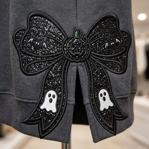 May include: A dark gray sweatshirt featuring a large, black glittery bow appliqu&eacute;. The bow is embroidered with a central jack-o'-lantern, two white ghosts on the bottom tails, and small bat silhouettes on the upper loops, all outlined in black thread.