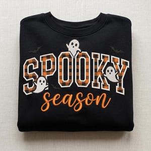 May include: A black crewneck sweatshirt featuring the embroidered text SPOOKY in an orange and black buffalo plaid pattern, with the word season in orange script below. Three white ghost figures and two small black bats are embroidered around.