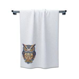 May include: White bath towel with a decorative owl design. The owl features intricate patterns in shades of blue, gold, and brown. The towel is hanging on a silver towel bar, perfect for a bathroom.