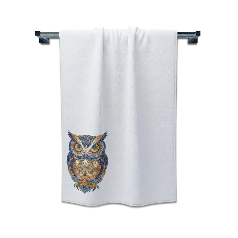 May include: White bath towel with a decorative owl design. The owl features intricate patterns in shades of blue, gold, and brown. The towel is hanging on a silver towel bar, perfect for a bathroom.
