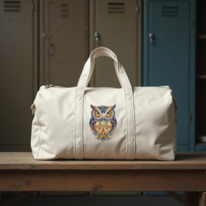 May include: Cream-colored duffel bag with a colorful owl design. The owl is depicted with blue, orange, and yellow details. The bag has two handles and a zipper closure, suitable for travel or gym use.