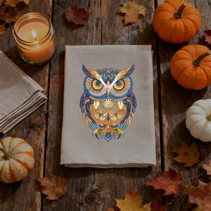 May include: A beige kitchen towel with a detailed owl design in blue, gold, and orange. The owl is centered on the towel, surrounded by pumpkins, fall leaves, and a lit candle on a wooden surface. A seasonal, decorative item.