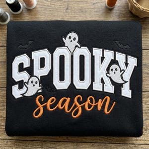 May include: A black sweatshirt featuring embroidered white block lettering that reads SPOOKY with three small white ghost figures and black bat silhouettes. Below, the word season is embroidered in orange cursive script lettering.