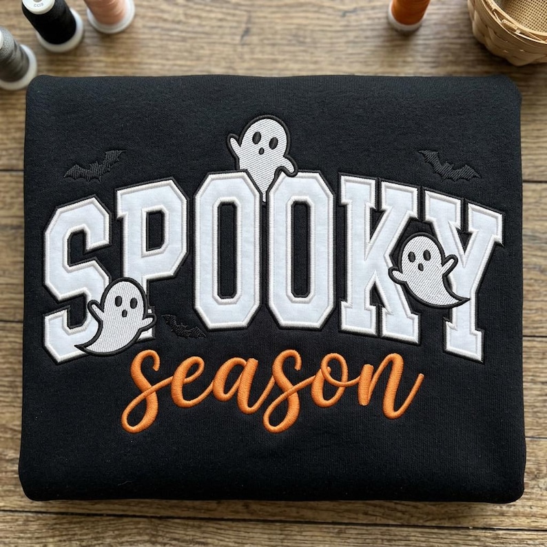 May include: A black sweatshirt featuring embroidered white block lettering that reads SPOOKY with three small white ghost figures and black bat silhouettes. Below, the word season is embroidered in orange cursive script lettering.