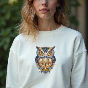 May include: Cream-colored sweatshirt featuring a detailed owl design. The owl is depicted with intricate patterns in shades of blue, gold, and orange. The sweatshirt has a classic crew neck and long sleeves, suitable for casual wear.