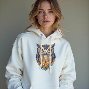 May include: Cream-colored hoodie featuring a detailed owl design on the front. The owl is rendered in shades of blue, orange, and yellow, with intricate patterns. The hoodie has a drawstring hood and a front pocket.