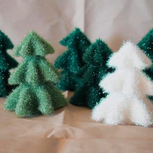 Op de afbeelding: Een verzameling kleine, decoratieve kerstbomen. De bomen zijn in tinten groen en wit, met een textuur en glitter. De bomen zijn gerangschikt op een lichtgekleurd oppervlak, wat een feestelijk vakantiethema suggereert.