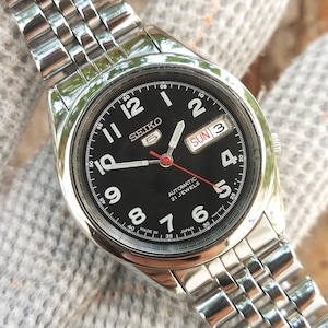 Puede incluir: Un reloj de pulsera automático Seiko 5 de acero inoxidable con esfera negra. El reloj presenta marcadores de hora blancos, una pantalla de día y fecha y un segundero rojo. La correa del reloj es de acero inoxidable.