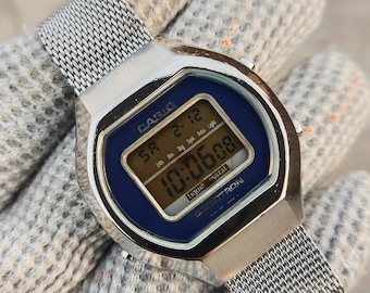 May include: A silver Casio digital watch with a blue face and a mesh band. The watch displays the time as 10:06:08. The watch face has the text "CASIO" and "LIQUID CRYSTAL".