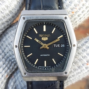 Może przedstawiać: Zegarek automatyczny Seiko 5 w stylu vintage z czarną tarczą i złotymi wskazówkami i znacznikami. Zegarek ma srebrną kopertę i czarny skórzany pasek. Dzień i data są wyświetlane o godzinie 3. Klasyczny zegarek.