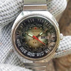 Może przedstawiać: Zegarek vintage Orient WD 21 Jewels ze srebrną bransoletą ze stali nierdzewnej. Tarcza zegarka ma zielono-czarną tarczę z wieloma strefami czasowymi, czerwoną wskazówką sekundową i wyświetlaczem daty. Zegarek ma obrotową ramkę.