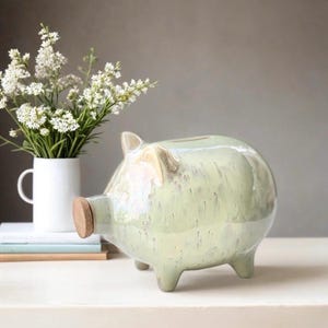 Könnte beinhalten: Ein hellgrünes Sparschwein aus Keramik mit einer Nase aus Kork und vier kleinen Beinen. Das Sparschwein hat einen Schlitz oben zum Einwerfen von Münzen. Links vom Sparschwein steht eine weiße Tasse mit weißen Blumen.