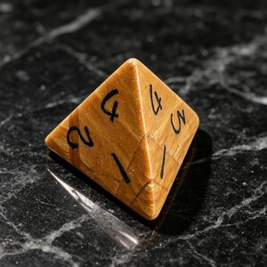 May include: A four-sided pyramid-shaped die crafted from a tan, wood-grained material. The faces of the die feature black ink numbers, including a visible 1, 2, 3, and 4, arranged in a standard numerical sequence for this type of gaming piece.