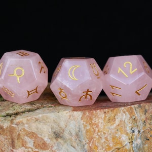 Puede incluir: Tres dados de piedra rosa con símbolos y números dorados. Los dados están facetados y presentan símbolos astrológicos y números. Los dados se muestran sobre una superficie texturizada, marrón y verde, sobre un fondo negro.