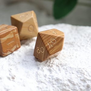 May include: Three polyhedral dice crafted from light brown wood with a distinct swirling grain pattern. Each die features engraved numbers filled with a metallic gold color. The dice are shaped as a cube, a diamond, and a multi-faceted geometric form.