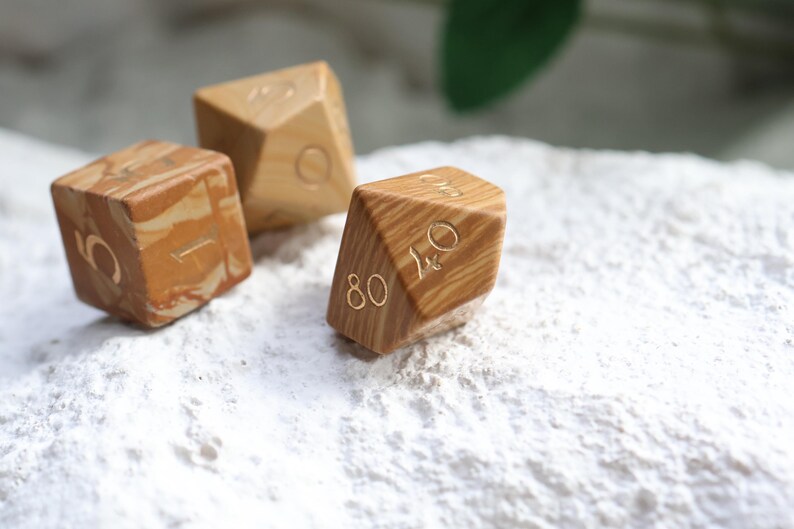 May include: Three polyhedral dice crafted from light brown wood with a distinct swirling grain pattern. Each die features engraved numbers filled with a metallic gold color. The dice are shaped as a cube, a diamond, and a multi-faceted geometric form.