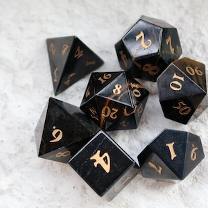 May include: A collection of seven black polyhedral dice with gold numbering. The dice are in various shapes, including a d4, d6, d8, d10, d12, and d20. The dice are displayed on a textured, off-white surface.