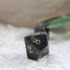 May include: Close-up of a black, multi-sided die with gold-colored numbers. The die is resting on a textured, white surface. The numbers 2, 8, and 0 are visible. The image suggests a game or tabletop activity.