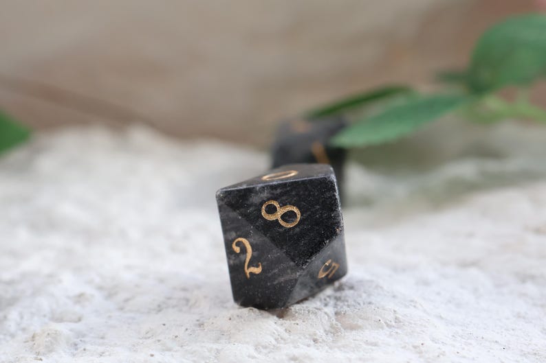 May include: Close-up of a black, multi-sided die with gold-colored numbers. The die is resting on a textured, white surface. The numbers 2, 8, and 0 are visible. The image suggests a game or tabletop activity.