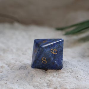 Op de afbeelding: Een donkerblauwe, veelzijdige achtzijdige dobbelsteen van steen met zichtbare natuurlijke donkere spikkels en goudkleurige gegraveerde cijfers. De dobbelsteen heeft een scherpe geometrische vorm met de cijfers acht, zes en twee duidelijk zichtbaar.