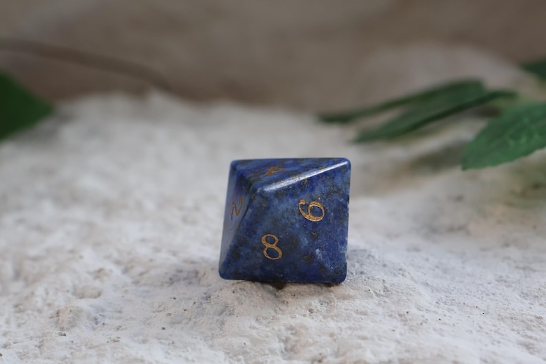 Op de afbeelding: Een donkerblauwe, veelzijdige achtzijdige dobbelsteen van steen met zichtbare natuurlijke donkere spikkels en goudkleurige gegraveerde cijfers. De dobbelsteen heeft een scherpe geometrische vorm met de cijfers acht, zes en twee duidelijk zichtbaar.