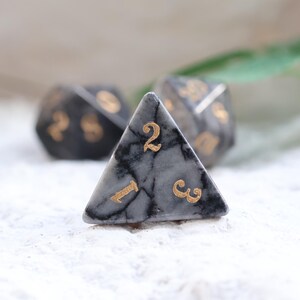 May include: Three polyhedral dice with a marbled gray and black pattern and gold numbering. The focus is on a triangular die with the number 2 visible. The other dice are out of focus.