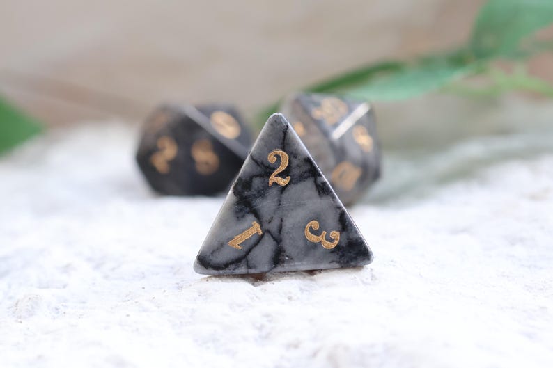 May include: Three polyhedral dice with a marbled gray and black pattern and gold numbering. The focus is on a triangular die with the number 2 visible. The other dice are out of focus.