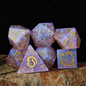Op de afbeelding: Een set van zeven polyhedrale dobbelstenen in lavendel- en blauwtinten met gouden cijfers en decoratieve accenten. De dobbelstenen liggen op een rots tegen een zwarte achtergrond. Voor rollenspellen.