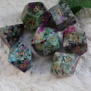 Może przedstawiać: Zestaw kości z kamieni szlachetnych w różnych kształtach, z zielonymi, fioletowymi i czarnymi odcieniami. Cyfry są wygrawerowane w kolorze złotym. Te kości są idealne do gier planszowych.