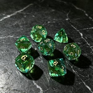Puede incluir: Un juego de siete dados poliédricos de color verde esmeralda translúcido con números dorados. La colección incluye un d20, d12, d10, d8, d6, d4 y un dado de porcentaje, todos con bordes afilados y una apariencia facetada similar a una gema.