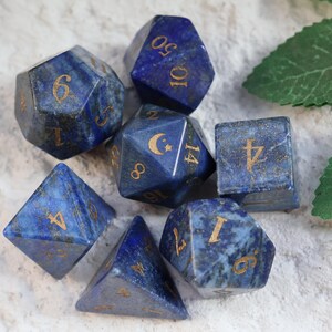Op de afbeelding: Een set van zeven veelzijdige dobbelstenen gemaakt van diepblauwe steen met goudkleurige cijfers. De dobbelstenen hebben verschillende geometrische vormen, waaronder een d20, d12, d10, d8, d6 en d4, met een maansikkel op een zijde van de d12.