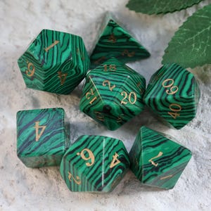 May include: Set of seven polyhedral dice in shades of green and black, with gold numbering. The dice have a marbled pattern and are arranged on a light-colored surface. Includes a D4, D6, D8, D10, D12, D20, and a percentile die.