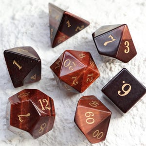 May include: A set of seven brown and red polyhedral dice with gold numerals, displayed on a textured white surface. The dice include various shapes, ideal for tabletop gaming, with measurements in centimetres.