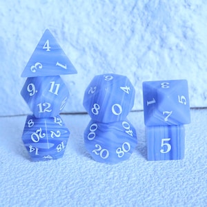 May include: A collection of blue and white marbled polyhedral dice for tabletop games. The set includes a four-sided die, a six-sided die, an eight-sided die, a ten-sided die, a twelve-sided die, a twenty-sided die, and a percentile die. Each die has white numbers.