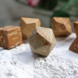 May include: A set of polyhedral dice crafted from tan and brown marbled stone. The dice feature engraved numbers filled with gold-colored ink. The central twenty-sided die displays the number 20 clearly, surrounded by other dice of various shapes.