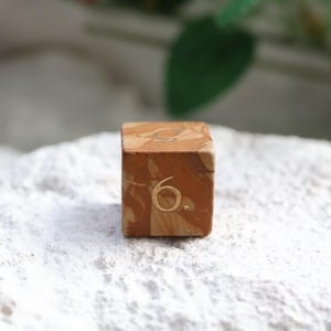 May include: A six-sided die featuring a marbled brown and tan pattern with the number six engraved in a light gold tone on the front face. The die has a smooth, cubic shape with rounded edges and a distinct, swirling color variation throughout.