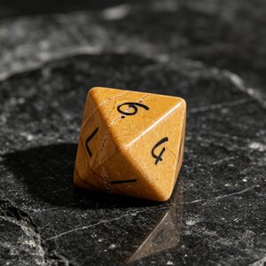 May include: An eight-sided die crafted from tan-colored stone with a natural veined texture. The die features black engraved numbers, including a clearly visible six and four, with sharp geometric edges and a polished, smooth surface finish.
