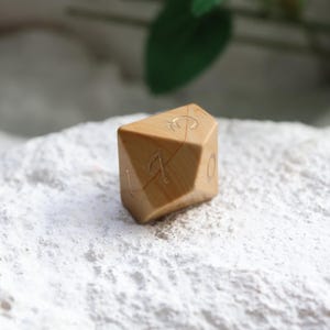 May include: A tan, multi-faceted polyhedral die with a smooth, polished finish and visible wood grain patterns. The die features engraved numbers, including a clearly visible 17 and 0, filled with a metallic gold color for contrast and readability.