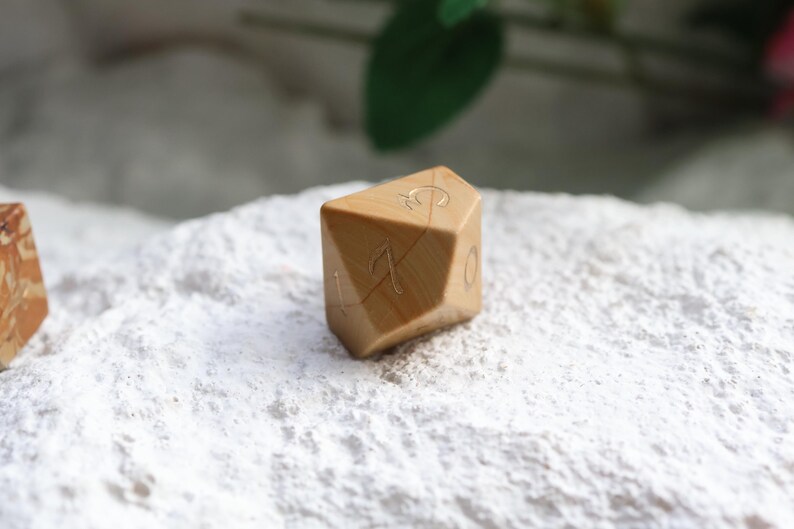 May include: A tan, multi-faceted polyhedral die with a smooth, polished finish and visible wood grain patterns. The die features engraved numbers, including a clearly visible 17 and 0, filled with a metallic gold color for contrast and readability.