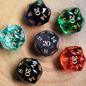 Peut inclure: Six dés à vingt faces multicolores, de couleurs variées : teal, vert, rouge, noir, bleu et bleu foncé avec des chiffres dorés. Les dés sont disposés sur une surface en bois brun clair.