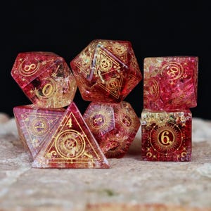 May include: Set of seven polyhedral dice in various shapes, including a d20, d12, d10, d8, d6, and d4. The dice are a translucent red with gold-colored designs and numbers. The dice are arranged on a textured surface against a black background.