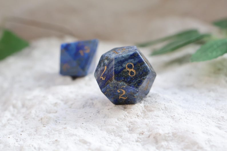 Op de afbeelding: Twee veelzijdige dobbelstenen gemaakt van diepblauwe lapis lazuli steen met natuurlijke goudkleurige spikkels. De voorste twintigzijdige dobbelsteen heeft gegraveerde goudkleurige cijfers, waaronder een acht en een twee, met een tweede dobbelsteen erachter.