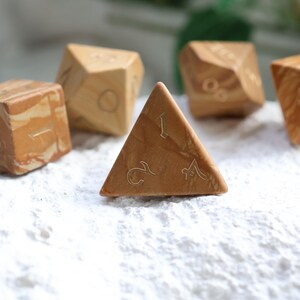 May include: A set of five polyhedral dice crafted from light brown and tan marbled material. Each die features engraved numbers in a thin, elegant font. The collection includes a four-sided triangular die, a cube, and other multi-faceted shapes.