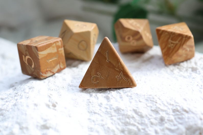 May include: A set of five polyhedral dice crafted from light brown and tan marbled material. Each die features engraved numbers in a thin, elegant font. The collection includes a four-sided triangular die, a cube, and other multi-faceted shapes.