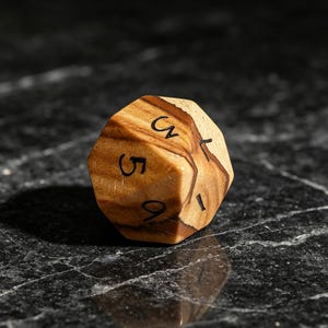 May include: A multi-faceted wooden die featuring a natural light and dark brown wood grain pattern. The die has black engraved numbers visible on its surface, including the digits 5 and 9, along with other partial numerical markings.