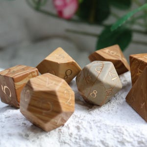 May include: A set of seven polyhedral dice crafted from light brown wood with visible grain patterns and engraved gold-colored numbers. The collection includes various geometric shapes such as cubes, pyramids, and icosahedrons, all featuring distinct numerical markings.