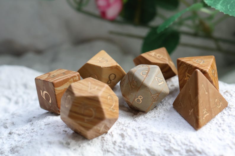 May include: A set of seven polyhedral dice crafted from light brown wood with visible grain patterns and engraved gold-colored numbers. The collection includes various geometric shapes such as cubes, pyramids, and icosahedrons, all featuring distinct numerical markings.